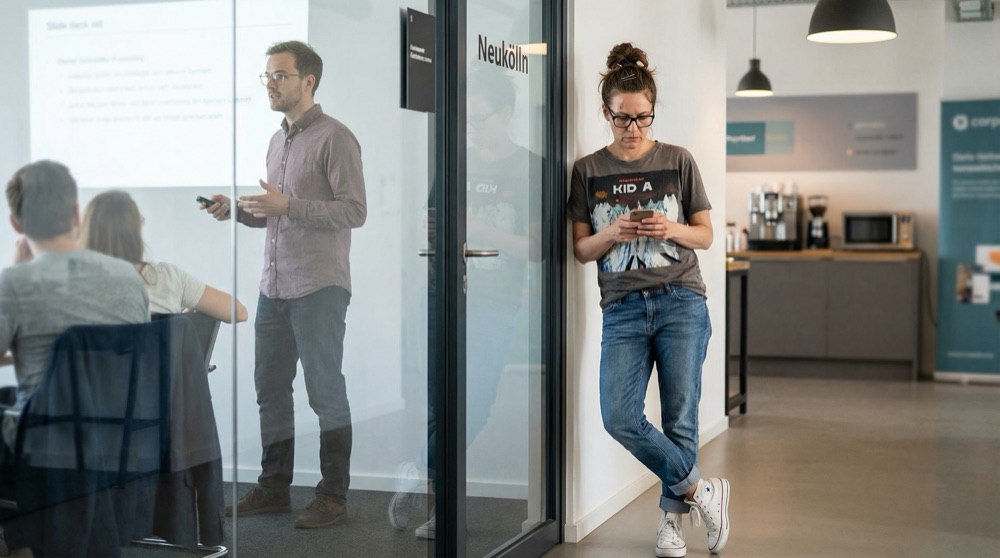 Katja standing in office hallway outside a glass conference room, gripping her phone with both hands, reading a new email notification, mid-morning office light